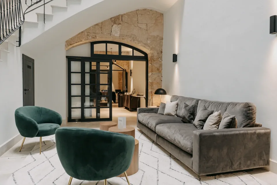 Zona de estar de CONVENDUM Palma con sillones verdes, sofá gris, escalera y un arco al fondo.