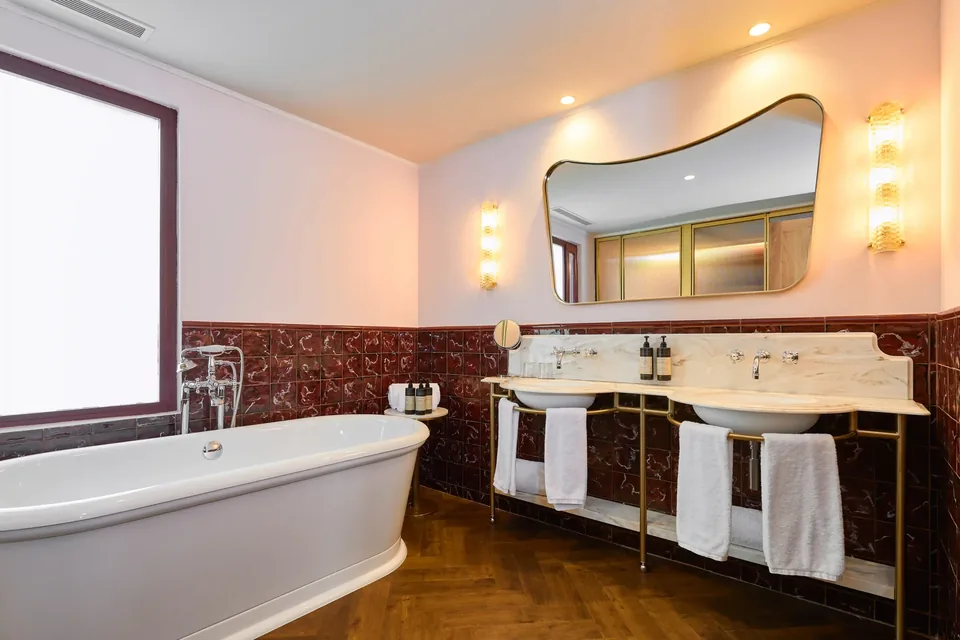 Baño del Hotel España Ocean Drive con bañera, dos lavabos, espejo y ventana junto a paredes claras y rojizas.