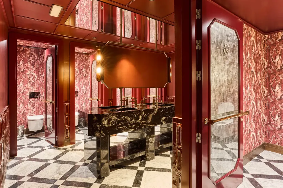 Baños del Hotel España Ocean Drive con lavabos oscuros, espejos, mamparas de tonos rojizos y pavimento geométrico.