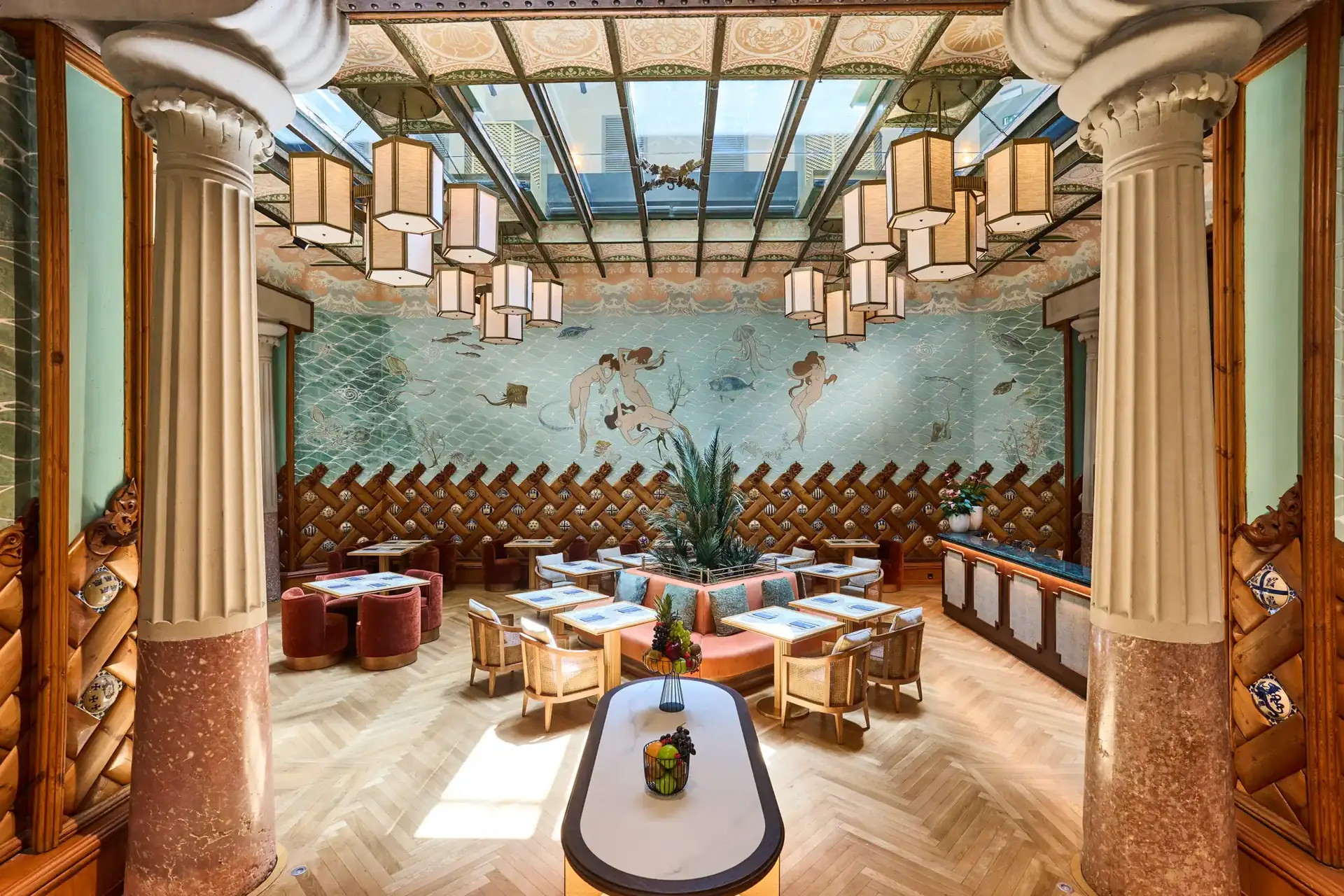 Sala del Hotel España Ocean Drive con columnas, cubierta acristalada, murales y asientos distribuidos alrededor de mesas.