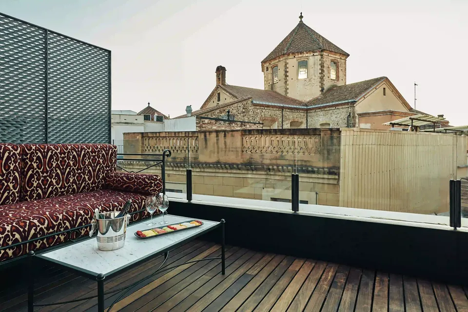 Terraza del Hotel España Ocean Drive con sofá, mesa baja y barandilla de vidrio frente a los tejados de Barcelona.