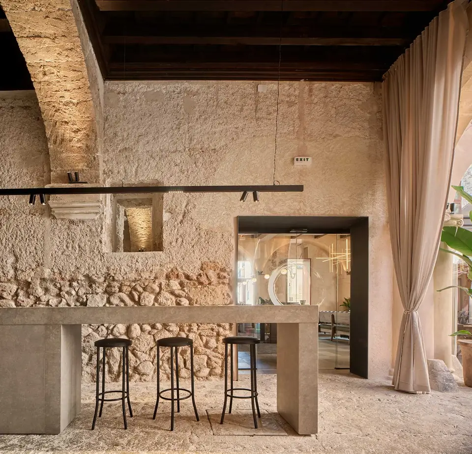 Barra de Nobis Hotel Palma con taburetes altos, muros de textura irregular y cortinas junto a una ventana.