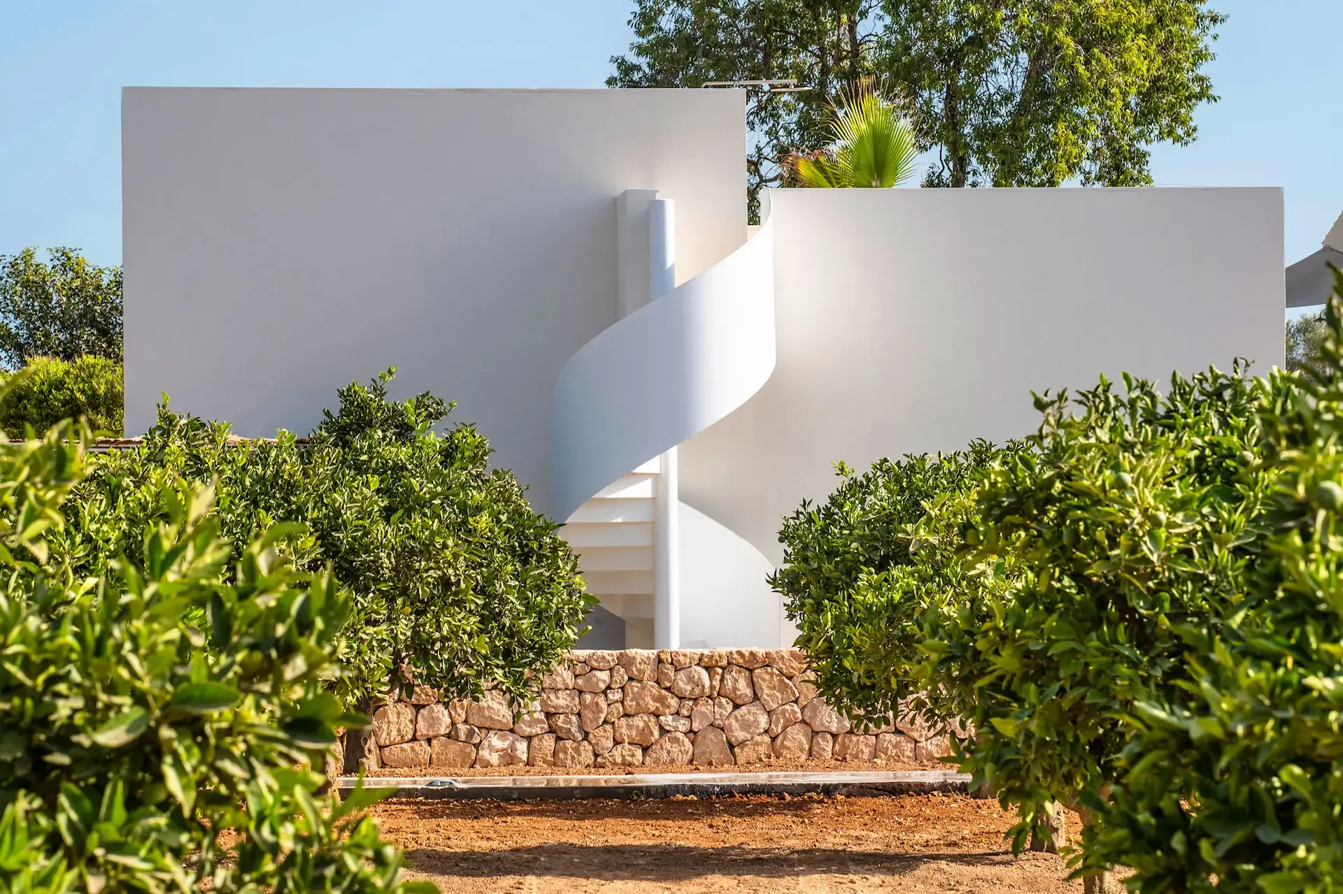 Exterior de OD Can Jaume en Ibiza con volúmenes blancos, escalera curva y árboles frente a un muro bajo.