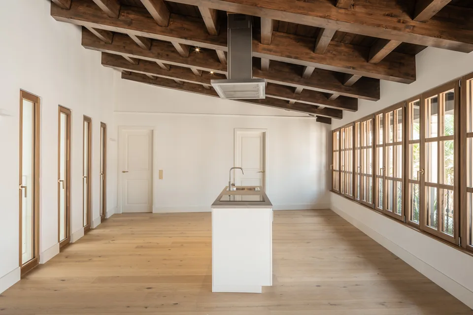 Cocina de la Carrer Sol en Palma con isla central, techo de vigas y una hilera de ventanas de marcos de tono madera.