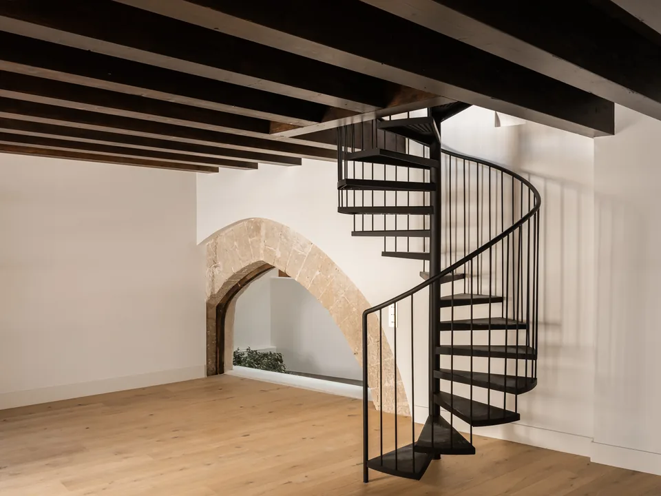 Sala de la Carrer Sol en Palma con escalera de caracol negra, techo de vigas y un hueco bajo un arco.