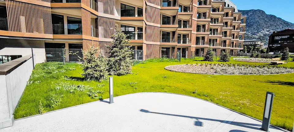 Fachada de TWA Andorra con balcones, paneles de tono madera y jardín con césped y árboles.