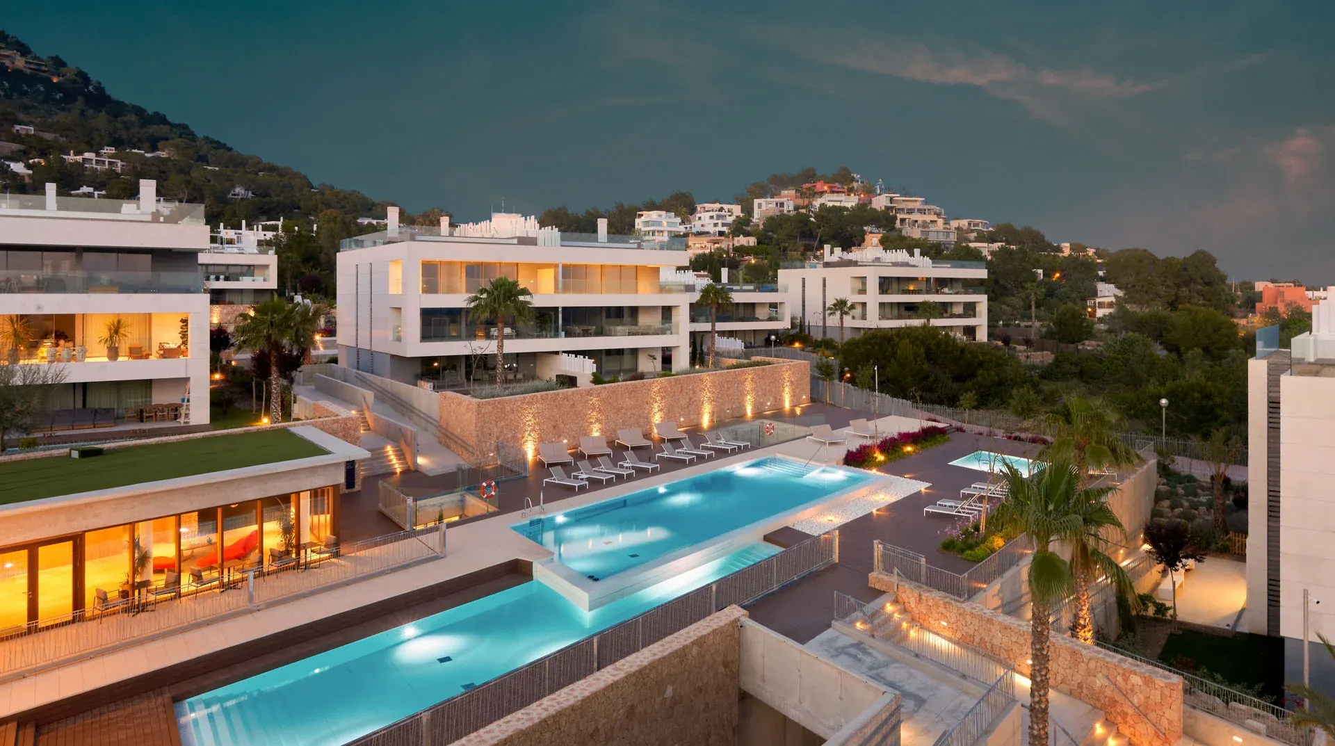 Vista elevada de TWA Talamanca con edificios claros, piscinas y terrazas iluminadas al anochecer.